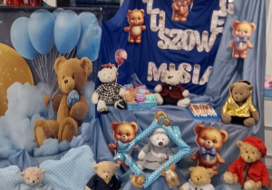 Dekoracja z okazji Dnia Pluszowego Misia: na granatowym materiale napis "Dzień Pluszowego Misia", na podłodze przykrytej błękitnym materiałem siedzą misie, a nad tablicą wisi balonowa girlanda w kolorze brązowo - błękitnym. Z jednej strony tablicy wisi misiowy baner.