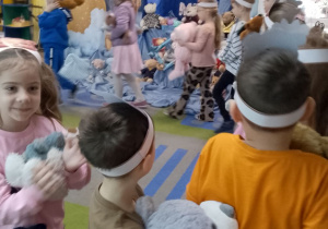 Przedszkolaki maszerują po kole ze swoimi misiami. W tle dekoracja z okazji Dnia Pluszowego Misia.