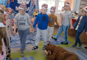 Dzieci idą w kole trzymając się za ręce, podczas zabawy "Stary niedźwiedź mocno śpi", a na środku koła leży nauczyciel w stroju misia.