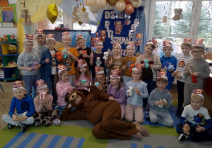 Zdjęcie grupowe dzieci z otrzymanymi upominkami i nauczyciela w stroju misia. Dzieci mają na głowach papierowe misiowe opaski.