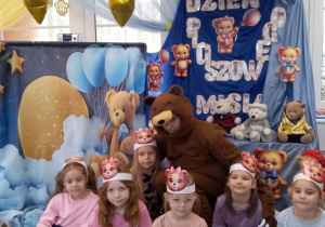 Grupka dziewczynek wspólnie z nauczycielem w stroju misia pozują na tle dekoracji.