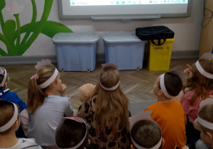 Dzieci siedzą na dywanie przed tablicą multimedialną i oglądają prezentację "Misie znane i nieznane".