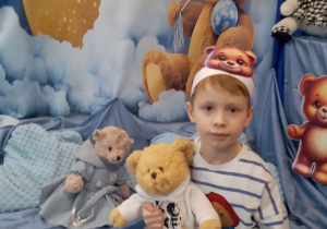 Adaś siedzi i trzyma misia na tle dekoracji.