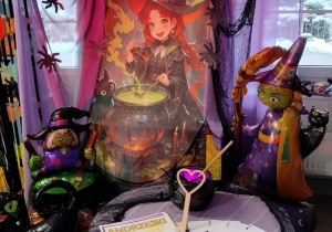 Dekoracja z okazji Andrzejek - na tablicy przypięty jest baner z czarownicą, po obu stronach banera wisi fioletowy materiał, na którym znajdują się papierowe pająki. Nad tablicą wiszą balonowe nietoperze. Obok tablicy stoją balonowe czarownice i kot. Przed tablicą stoi stolik nakryty fioletowym materiałem, na którym stoi kociołek i leżą karty i klucz.