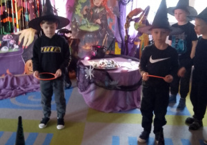 Na tle dekoracji stoją chłopcy i rzucają obręczami na balonowe czarne kapelusze.