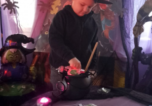 Chłopiec w czarnym kapeluszu na głowie losuje z czarnego kociołka ciasteczkową wróżbę na tle dekoracji.