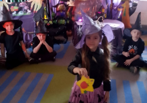 Przedszkolaki siedzą na dywanie, a w środku koła leżą żółte gwiazdki z wróżbami i czerwony kręgiel. Dziewczynka w kapeluszu na głowie prezentuje wylosowaną wróżbę. W tle dekoracja z okazji Andrzejek.