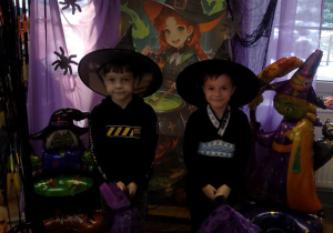 Dwóch chłopców w kapeluszach na głowie pozuje do zdjęcia na tle dekoracji z miotłami.