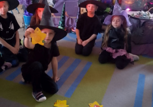 Dzieci siedzą na dywanie, a w środku koła leżą żółte gwiazdki z wróżbami i czerwony kręgiel. Chłopiec w kapeluszu na głowie prezentuje wylosowaną wróżbę. W tle dekoracja z okazji Andrzejek.