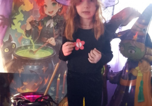 Dziewczynka w kapeluszu na głowie prezentuje wylosowaną wróżbę w kształcie kwiatka. W tle dekoracja z okazji Andrzejek.