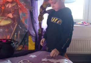 Chłopiec w kapeluszu na głowie losuje wróżbę w kształcie kwiatka. W tle dekoracja z okazji Andrzejek.