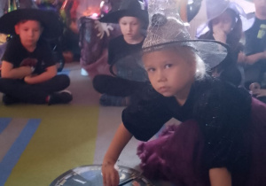 "Słoneczka" siedzą w kole na dywanie, a w środku dziewczynka kręci kołem fortuny z wróżbami.