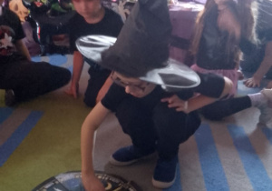 Dzieci siedzą w kole na dywanie, a w środku chłopiec w czarnym kapeluszu kręci kołem fortuny z wróżbami.