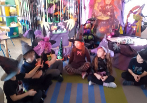 "Słoneczka" siedzą w kole na dywanie i podają sobie czarodziejską miotłę. W tle dekoracja z okazji Andrzejek.