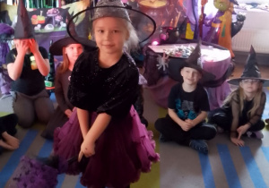 Dzieci siedzą w kole na dywanie, a w środku dziewczynka tańczy z miotłą.