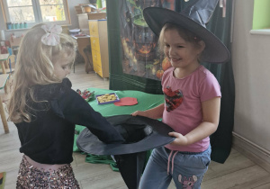 Klara w stroju czarownicy trzyma kapelusz wiedźmy, z którego Alicja losuje wróżbę.