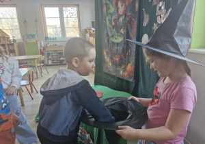 Klara w kapeluszu czarownicy na głowie trzyma w dłoniach kapelusz z wróżbami. Swoją wróżbę losuje Szymon.