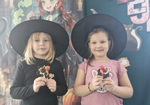 Nadia i Maja w kapeluszach czarownic pozują do zdjęcia na tle andrzejkowej dekoracji.