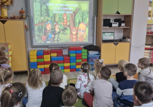 Dzieci siedzą na dywanie i oglądają film edukacyjny.