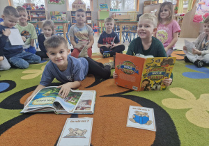 Staś i Lucjan z książkami w dłoniach siedzą na dywanie. Przed chłopcami leża karty z zabawy "Naśladuj misia".