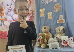 Wojtuś na tle dekoracji stoi trzymając w dłoniach lupę oraz kartę przedstawiającą misia z lupą.