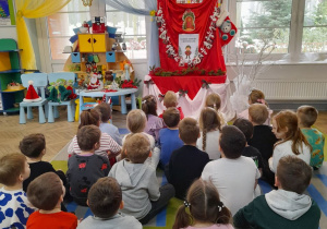 Dzieci siedzą na dywanie, a przed nimi na tablicy znajduje się kalendarz adwentowy. Obok na stoliku leżą ozdoby świąteczne.