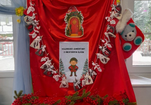 Na tablicy przykrytej czerwonym i białym materiałem przypięty jest kalendarz adwentowy z niesfornym Elfem. Nad tablicą znajduje się Elf, który trzyma list do dzieci.