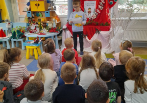 Dzieci siedzą na dywanie, a przed nimi stoi Mikołaj i czyta dzieciom list od Elfa.