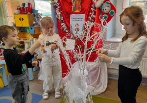 Na dywanie stoi wazon z białymi gałązkami. Troje dzieci wiesza na gałęziach papierowe aniołki. W tle tablica z kalendarzem adwentowym.