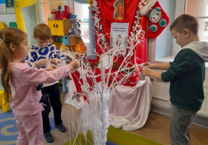 Marcel, Adam i Amanda wieszają na białych gałęziach umieszczonych w wazonie papierowe aniołki. W tle tablica z kalendarzem adwentowym.