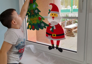 Jakub wiesza na oknie obok Mikołaja choinkę z napisem "Wesołych Świąt".