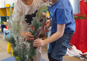 Staś i Tosia wkładają świąteczne gałązki do wazonu, który stoi na dywanie.