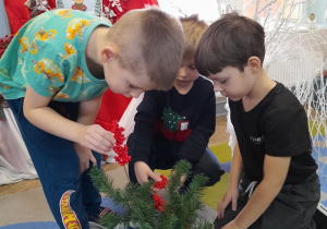 Trzech chłopców układa w wazonie kompozycję z gałązek iglastych i gałązek z czerwonymi kulkami. W tle kalendarz adwentowy.