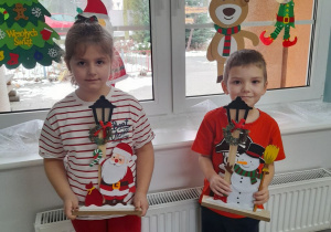 Nadia i Jakub stoją na tle okna i trzymają dekoracje świąteczne: Mikołaja i bałwanka.