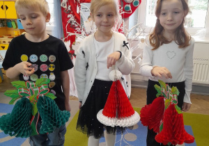 Fabian, Nikola i Emma stoją obok siebie. Dzieci trzymają świąteczne ozdoby z papieru: dzwonki w kolorze zielonym i czerwonym oraz czapkę Mikołaja.