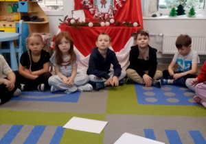 Dzieci siedzą na dywanie w półkolu. Przed nimi leżą odwrócone obrazki i kartoniki z cyframi. W tle tablica z kalendarzem adwentowym.