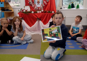 Dzieci siedzą na dywanie. W środku koła siedzi Antek, który trzyma odsłoniętą ilustrację.