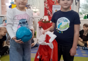 Na dywanie stoi Marysia trzymająca piłkę i Marcel z workiem.