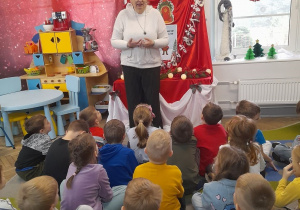Dzieci siedzą na dywanie, a przed nimi stoi Pani z biblioteki. Zaproszony gość opowiada o Świętym Mikołaju. W tle tablica z kalendarzem adwentowym, kącik domowy.