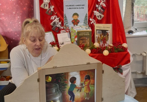 Pani z biblioteki czyta bajkę pt. "Prezenty Świętego Mikołaja" prezentując ją w formie Teatrzyku Kamishibai. W tle tablica z kalendarzem adwentowym.