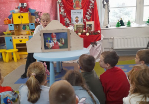 Pani z biblioteki siedzi przy stoliku, na którym stoi Teatrzyk Kamishibai. Dzieci słuchają bajki czytanej przez zaproszonego gościa pt. "Prezenty Świętego Mikołaja".