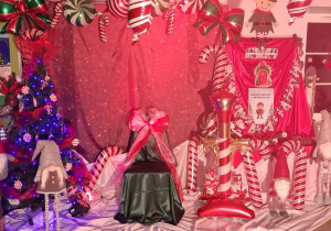 Dekoracja mikołajkowa: na tle czerwonego banera stoi krzesło okryte zielonym materiałem i przewiązaną wstążką biało-czerwoną , nad banerem wisi girlanda balonowa w kolorze czerwono-biało-zielonym. Obok krzesła stoi choinka, krasnale i balonowe świąteczne dekoracje.