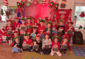 Zdjęcie grupowe dzieci w mikołajkowych opaskach na tle dekoracji z otrzymanymi upominkami ufundowanymi przez rodziców z grupy.