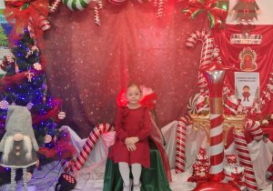 Zuzia siedzi na krześle wyłożonym zielonym materiałem. W tle mikołajkowa dekoracja.