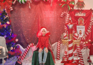 Chłopiec w czapce Mikołaja siedzi na krześle wyłożonym zielonym materiałem na tle dekoracji.