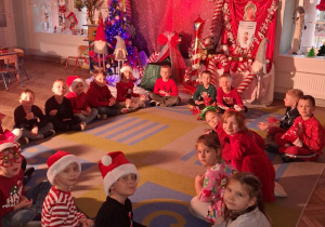 Przedszkolaki siedzą na dywanie na tle dekoracji.