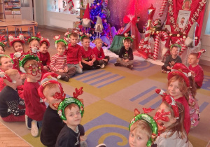 Dzieci w mikołajkowych opaskach siedzą na dywanie na tle dekoracji.