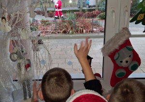 "Słoneczka" stoją przy oknie, przez które widać idącego Mikołaja.