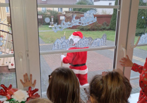 Grupka dzieci stoi przy oknie, przez które widać idącego Mikołaja.