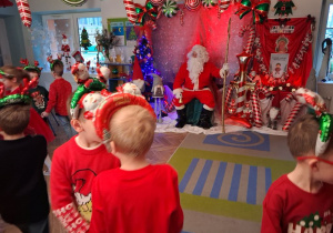 "Słoneczka" ustawione w małych kółeczkach śpiewają świąteczną piosenkę przed Mikołajem, który siedzi na krześle.
